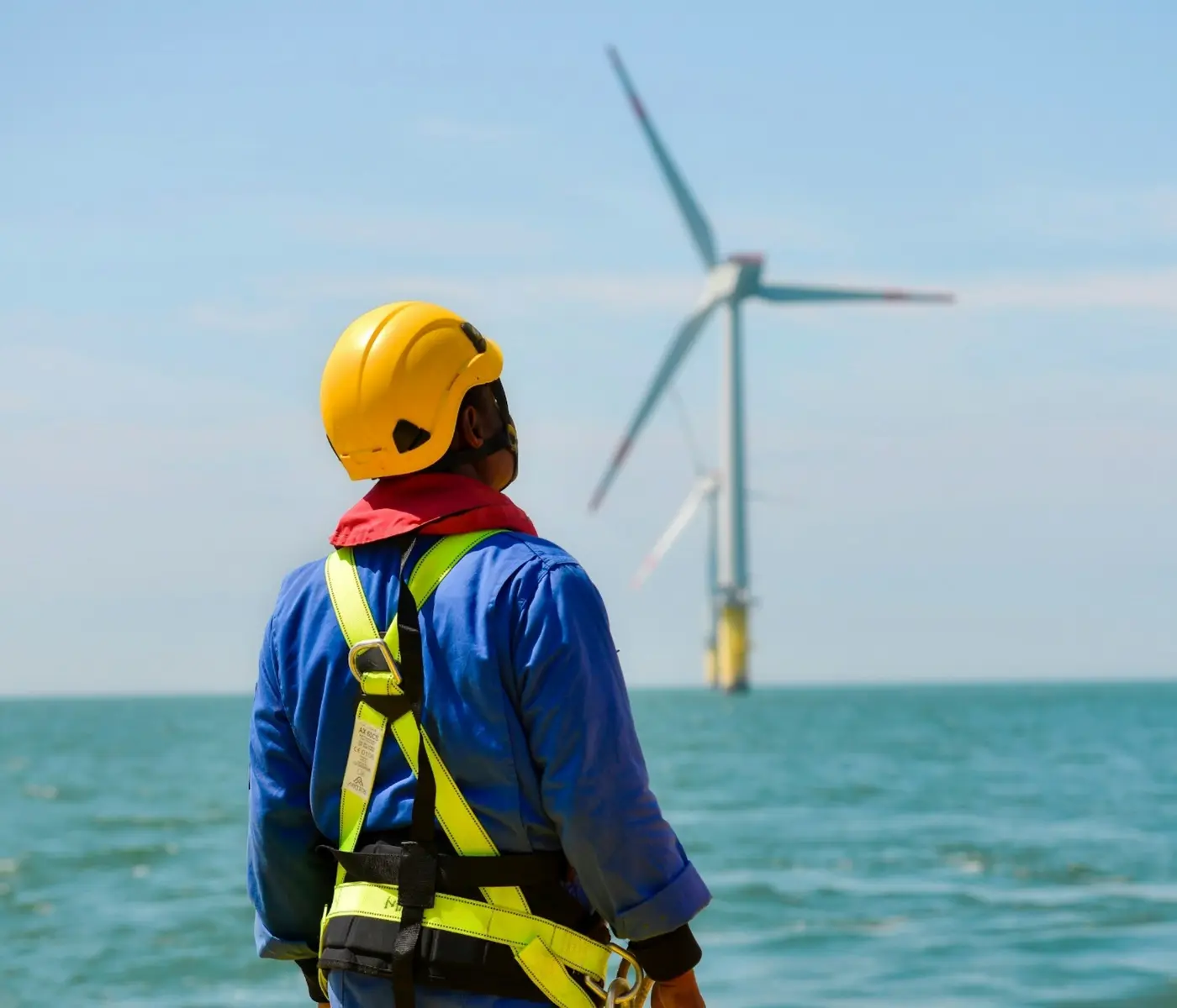 A technician in a hard hat and harness looking out at an offshore wind turbine.