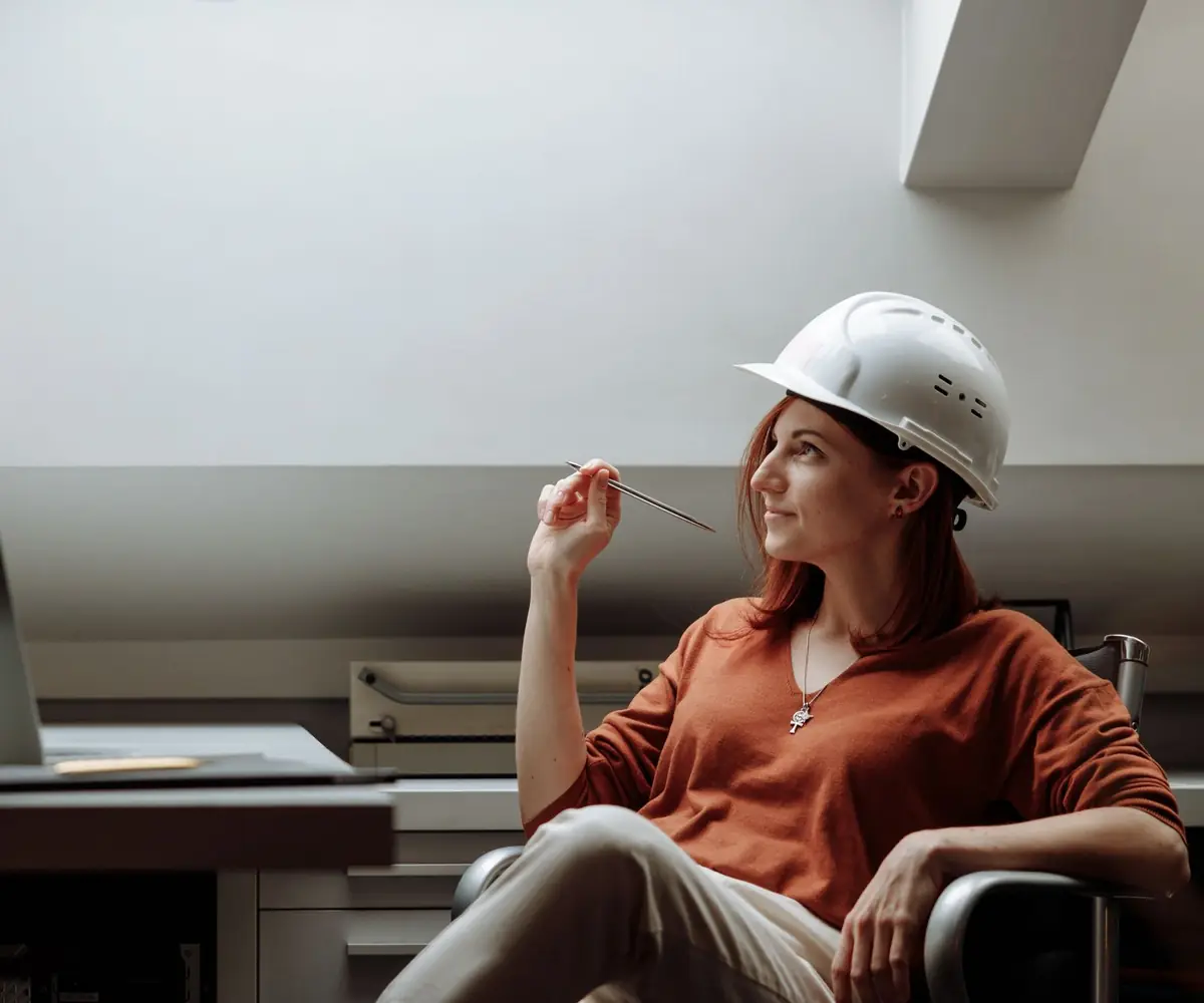 An engineer in a hard hat sitting back at her desk, thinking something through.